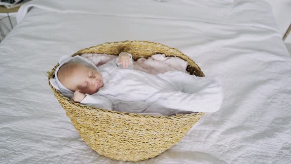Newborn Baby in White Bodysuit Sleeps in Small Brown Basket