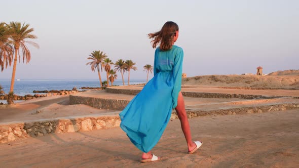 Young Woman in Blue Flying Dress is Walking By the Sea Sand Coast at Sunset