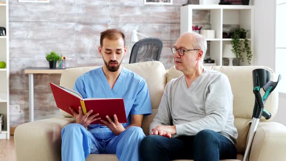 Social Worker Reading a Book To an Old Disabled Man in Nursing Home alt