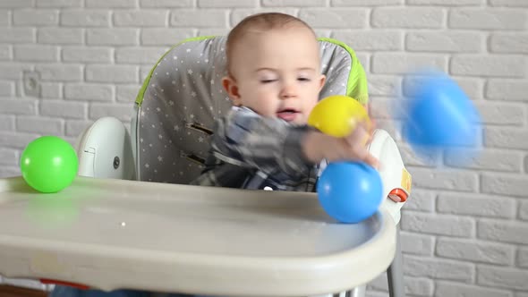 Baby Boy in a Plaid Shirt Sitting at a Children's Table Throws Multicolored Plastic Balls alt