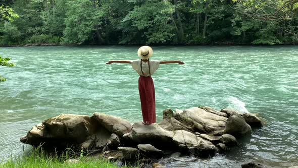 Young beautiful vintage woman in straw boater hat raising hands up near mountain river travel nature