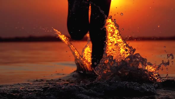 Silhouette of Legs of Boy Running Into the Water at Sunset and Creating Splashes. Slow Motion alt