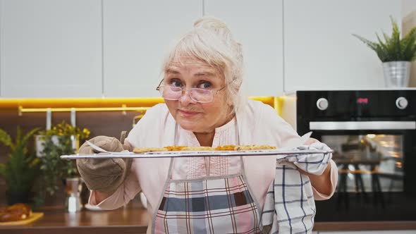 Grandmother in Apron is Smelling Hot Cookies on Baking Sheet and Smiling Being Satisfied Cooking at alt