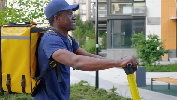 Smiling Afroamerican Man Courier Delivery Walks Street with Electric Scooter alt