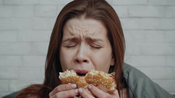 Unhappy Young Woman Eating Burger Looking At Camera In Bedroom alt