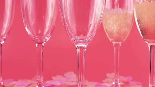 Sparkling wine is poured into glasses on a pink background with heart-shaped confetti. alt