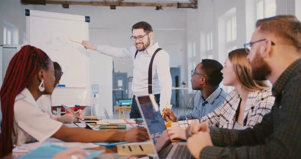 Happy Male Financial Company Boss Working on Flipchart Diagram with Colleagues During Team Meeting alt