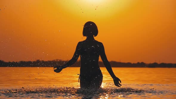 Silhouette of Woman at Sunset Raises Hands Up and Creating Splashes of Water alt