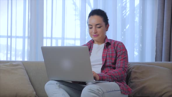Young Serious Woman Freelancer Working on a Laptop Sitting on a Sofa at Home, Businesswoman Typing alt