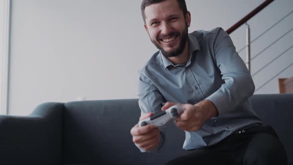 Happy Young Man Is Sitting on Sofa and Enjoying New Video Game Holding Joystick Controller
