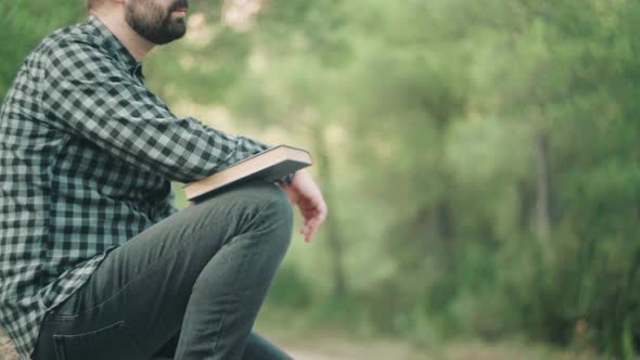 Caucasian Man With Book Sitting On The Ground With Green Nature In The Background. - close up alt