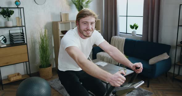 Guy in Training Clothes which Exercising on Stationary Bike and Looking Into Camera alt