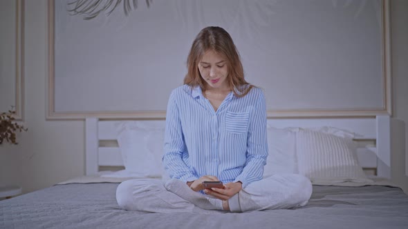Female Wearing Pajamas Typing Message on Mobile in Bedroom