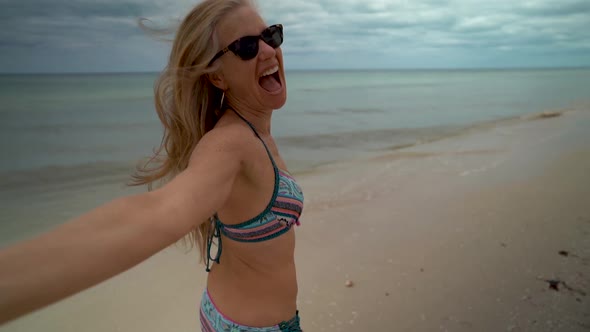 Slow motion with flying hair, a playful, mature woman in sunglasses and bikini running along the bea alt