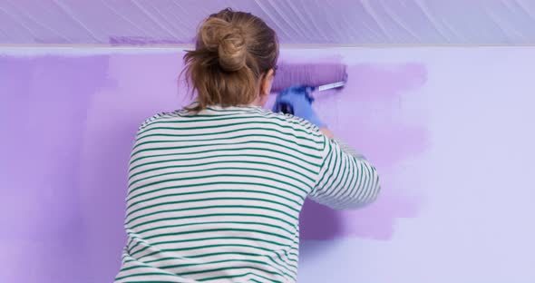 Young Woman in Glasses and Working Rubber Gloves Repaints Wall in Room with Roller in Brighter Shade