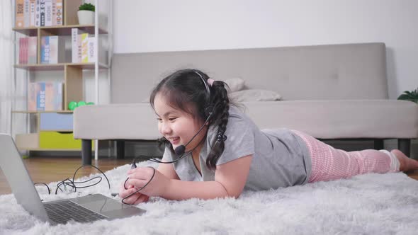 Asian girl lying and taking video call on laptop with wireless microphone in living room