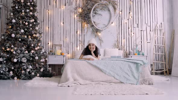 Happy Happy Young Lady in Winter Clothes with Hat Lying on the Bed. alt