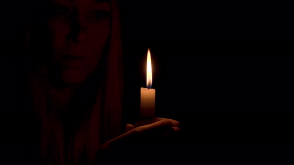A burning candle illuminates the face of a beautiful young woman in the dark. alt
