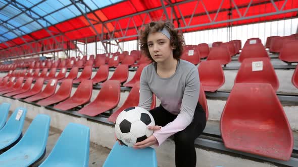 Young serious girl with soccer ball sitting on seats at football stadium alt