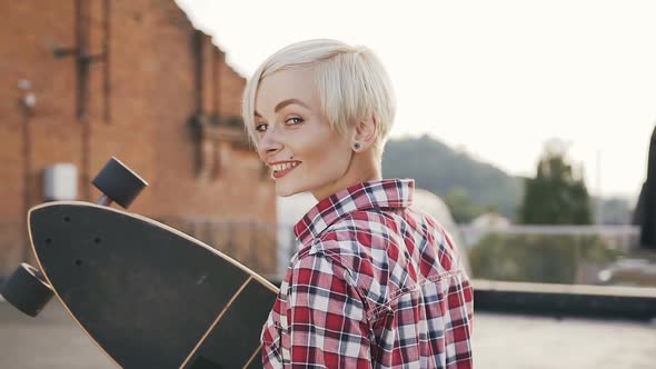 Beautiful Blonde Girl Walking with Long Skateboard on the Modern Building Roof alt