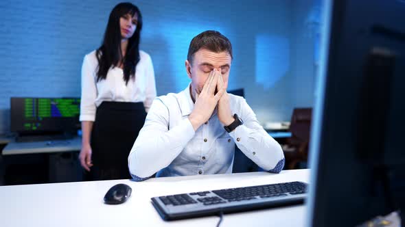Exhausted Man Rubbing Face As Nervous Woman Yelling and Gesturing at Background in Blue Office alt