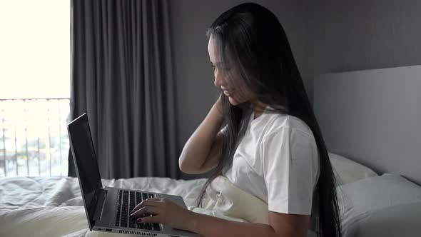 A young woman sitting on a bed in room and works on their laptop.