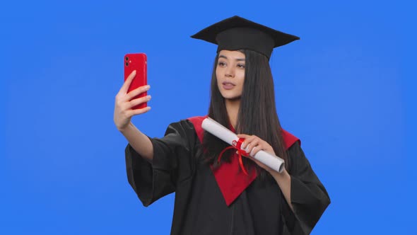 Portrait of Female Student in Graduation Costume Holding Diploma and Making Selfie on Mobile Phone alt