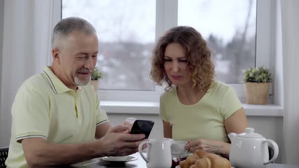 A Married Couple Watches the News on the Phone Over Breakfast in the Kitchen