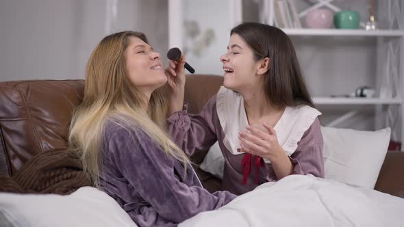 Joyful Pretty Woman Laughing Applying Face Powder Resting with Friend at Pajama Party Indoors alt