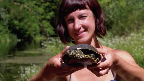 Woman Holds Funny Turtle in Arm and Smiles on River with Green Vegetation alt