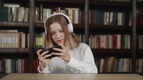 Lazy Schoolgirl with White Headphones Smartphone Distracting From Homework Sitting in Library alt