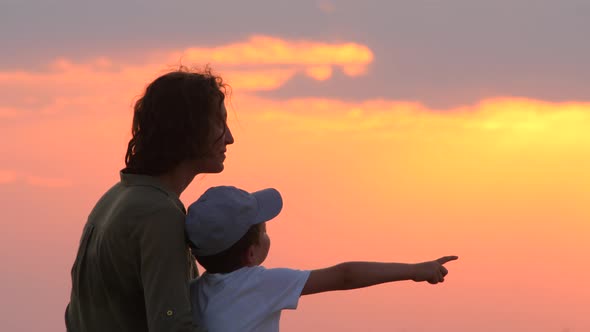 Beautiful woman hugging her kid watching amazing sunrise over the sea, city. Happy childhood concept