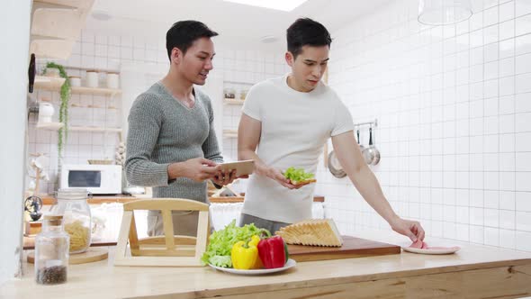 Asia gay couple using tablet and preparing the breakfast, sandwich vegetable on table
