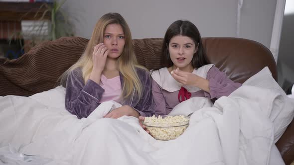 Two Absorbed Young Caucasian Women in Pajamas Eating Popcorn Looking at Camera Watching Film on TV alt