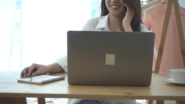 young smiling asian woman working on laptop while sitting in a living room at home using phone. alt