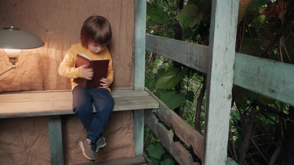 Cute Little Child on the Porch of a Village House is Trying to Read a Book alt