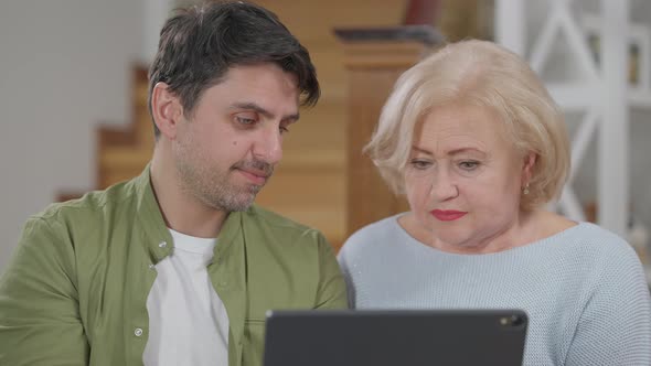 Portrait of Confused Senior Woman and Patient Adult Man Talking Pointing at Tablet alt