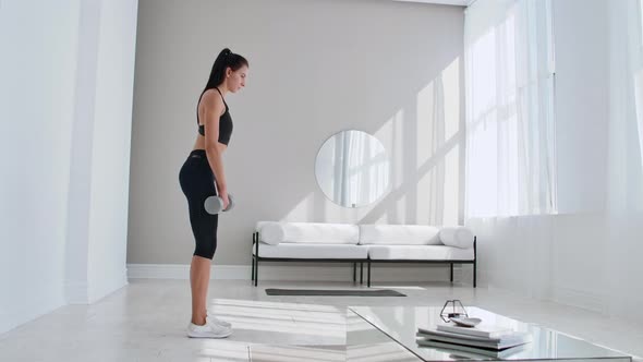The Beautiful Female Performs Leans Forward with Dumbbells in Her Hands in the White Interior of the alt
