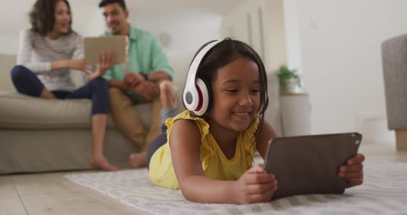 Happy hispanic girl watching video on tablet with parents sitting on sofa in background alt