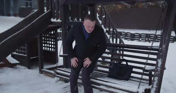 An Adult Man Puts His Briefcase And Sits On A Wooden Swing In A Snowy Yard
