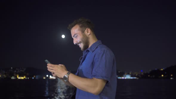 Young man using phone at night against city view by the sea. alt
