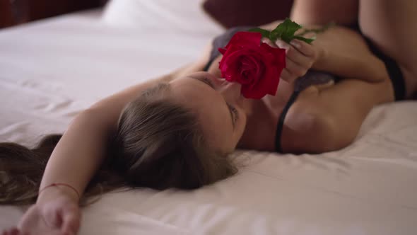 Young Slim Woman Lying on Comfortable Bed in Lingerie Smelling Red Rose in Slow Motion Smiling alt