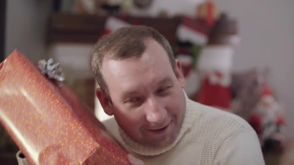 Closeup Portrait of Excited Adult Caucasian Man Shaking Christmas Gift Box and Smiling alt