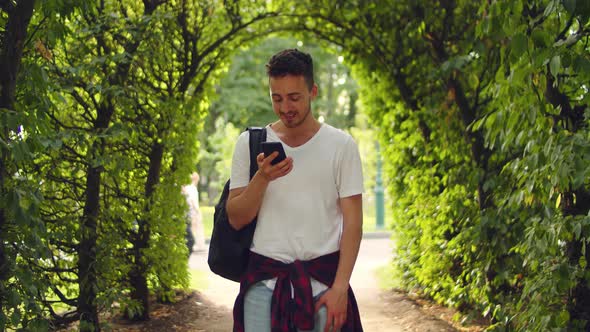 Handsome Young Man Stands in the Park Inside a Tunnel of Trees with a Phone in His Hands alt