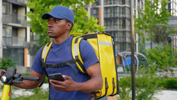 Afroamerican Man Courier Walks with Electric Scooter Uses Smartphone Navigate alt