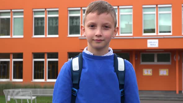 A Young Boy Smiles and Waves at the Camera - an Elementary School in the Background alt