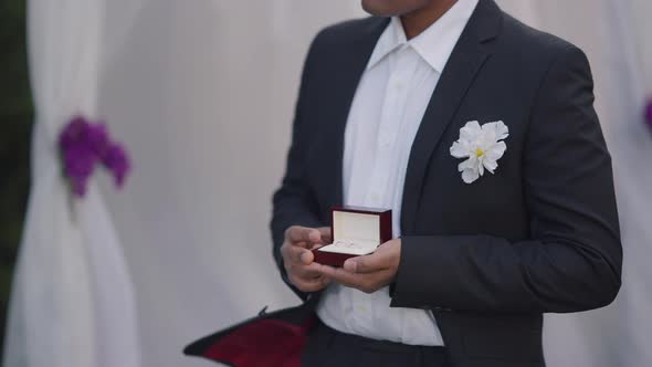 Unrecognizable African American Man in Suit with Boutonniere Holding Wedding Rings Box Standing at alt
