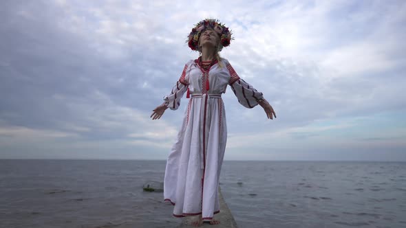 Young Calm Confident Ukrainian Woman in Embroidered Dress Standing on River Pier Raising Hands in alt