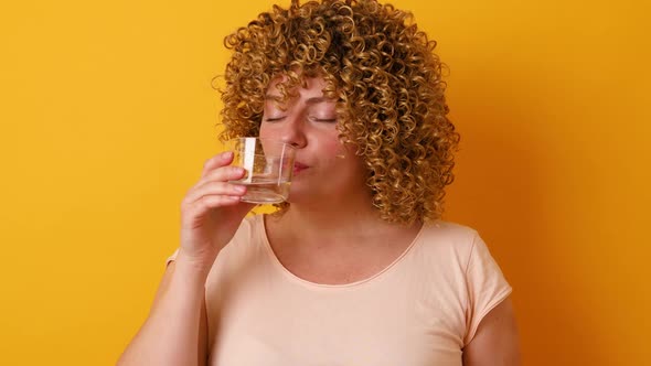 Beautiful Young Woman with Curly Fluffy Hair Drinking Glass of Water Over Yellow Background