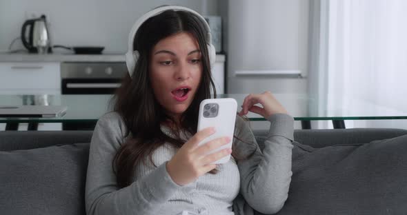 A Shocked Brunette Girl in a Grey Sweater Saying Wow Looking Smartphone and Covering Her Mouth While alt